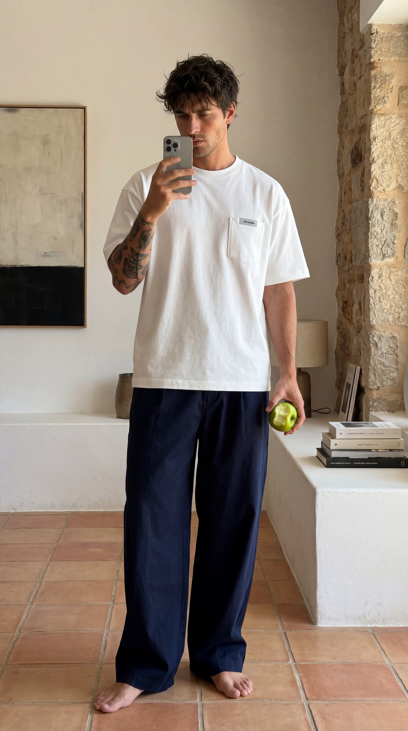Man taking a selfie in a white JAN KORBA t-shirt and navy pants in a room with stone walls.